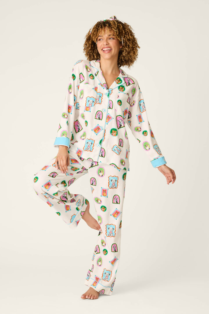 A smiling person with curly hair stands on one foot against a plain, light background, wearing the P.J. Salvage Picture Purrfect Pet Portraits 3-Piece Flannel PJ Set. The pajamas feature vibrant, cartoon-inspired prints with blue trim at the cuffs and collar.