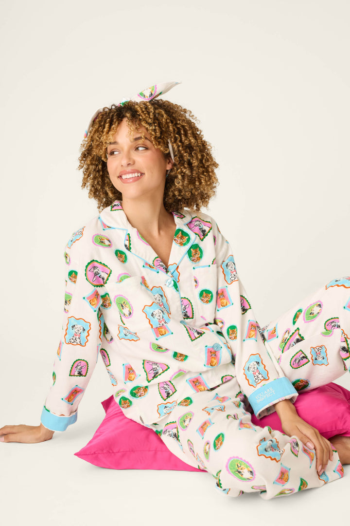 A woman with curly hair, smiling and looking to the side, sits on a bright pink pillow against a plain background. She wears the Picture Purrfect Pet Portraits 3-Piece Flannel PJ Set by P.J. Salvage, featuring colorful cartoon prints and a matching headband.
