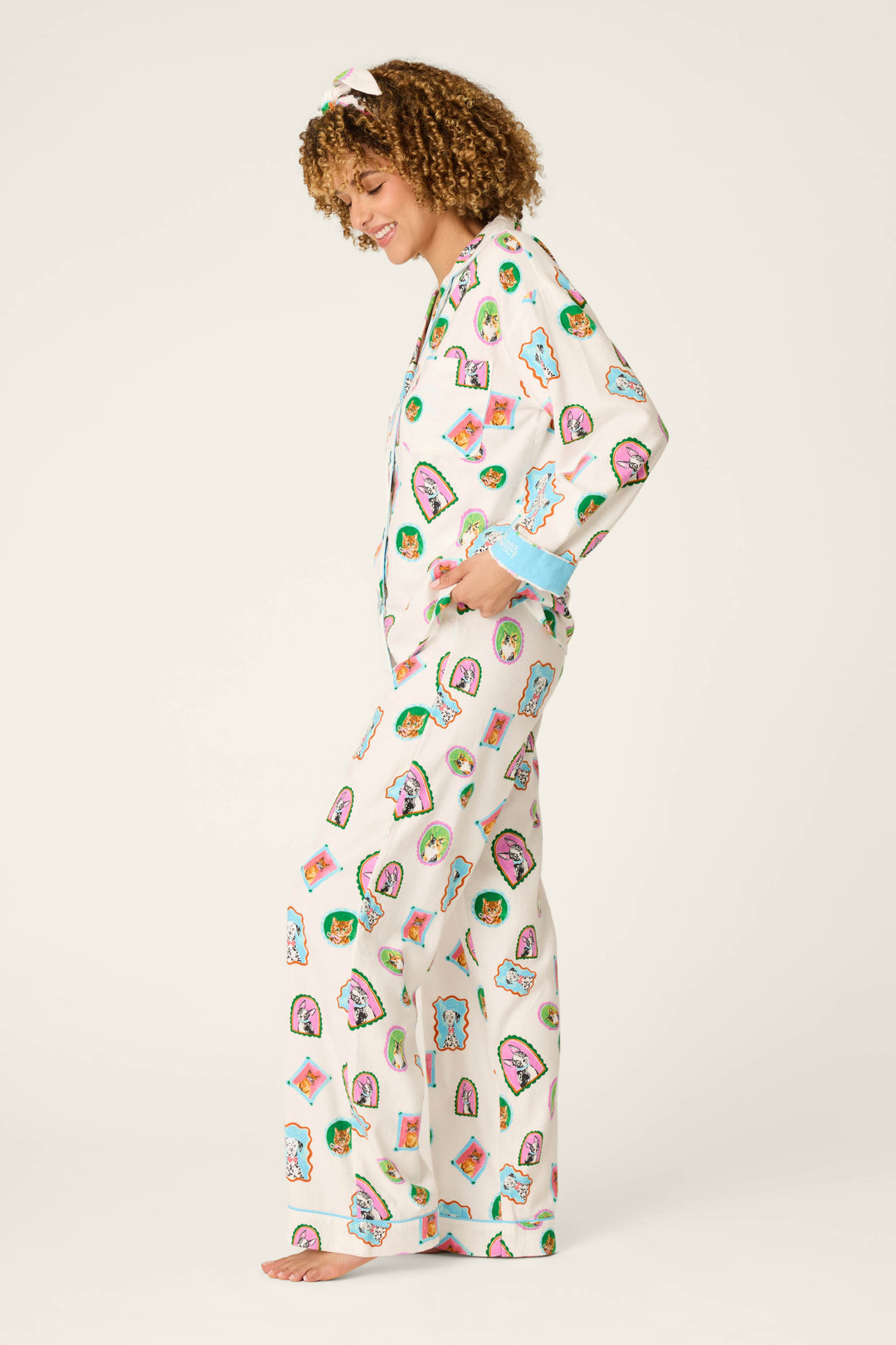 Wearing the P.J. Salvage Picture Purrfect Pet Portraits 3-Piece Flannel PJ Set, a person with curly hair stands barefoot in profile, smiling in white pajamas with playful taco and avocado prints. The pajama top features blue cuffs and is paired with a matching headband.