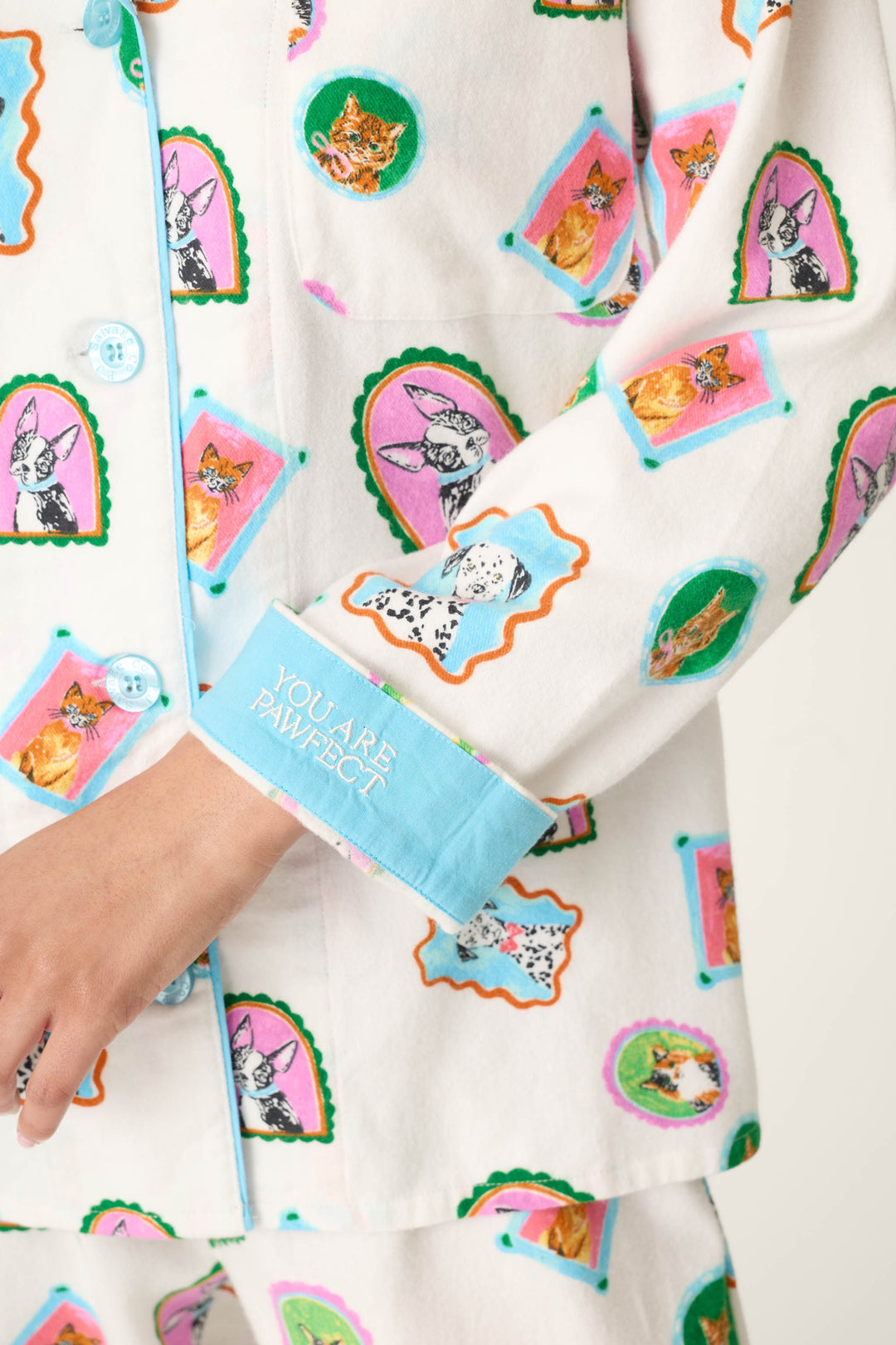 A person is wearing the P.J. Salvage Picture Purrfect Pet Portraits 3-Piece Flannel PJ Set, which features white fabric decorated with colorful cat and dog portraits and blue trim. The sleeve cuff displays "YOU ARE PAWFECT" in white text. Only part of the person's hand is visible.