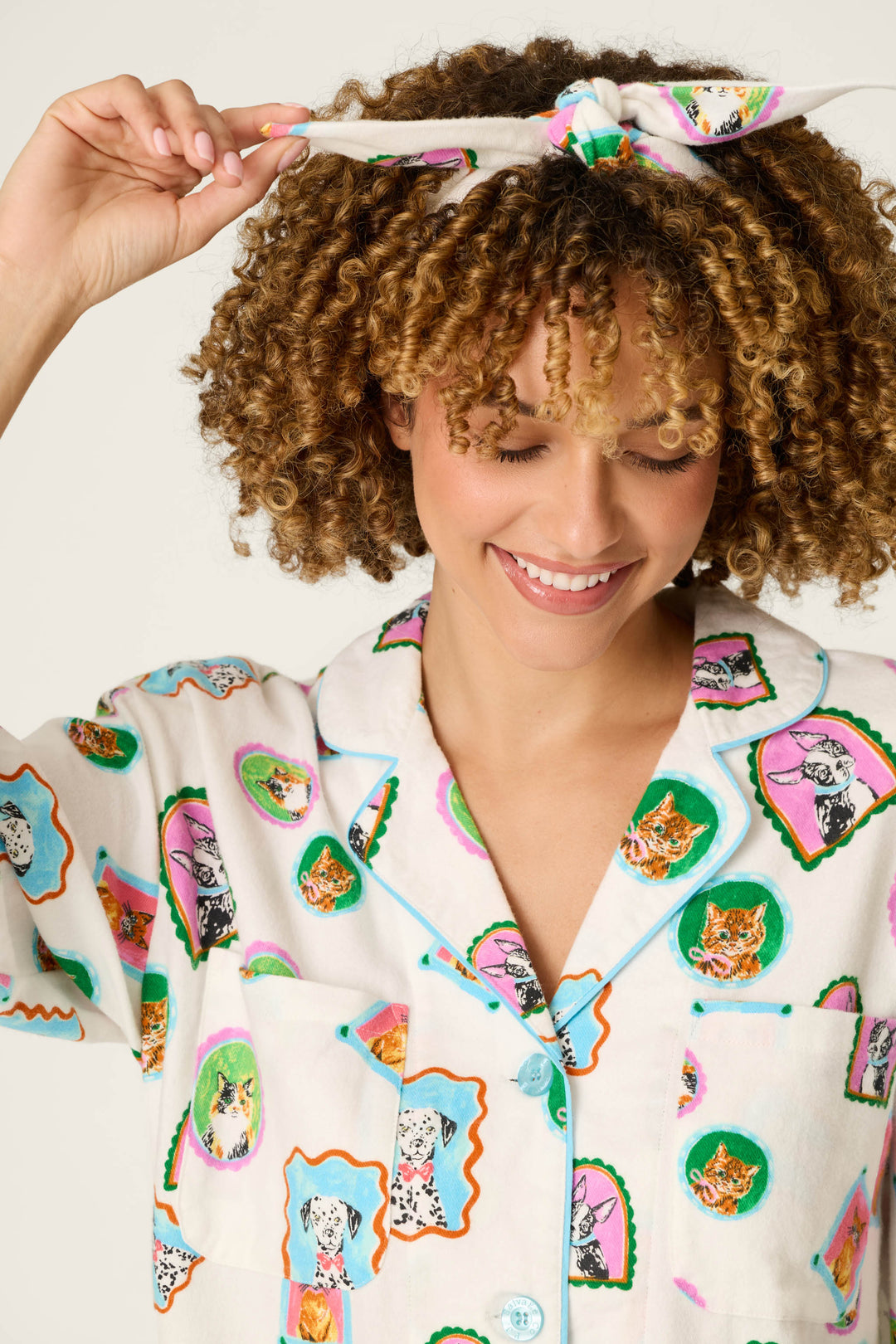 A smiling person with curly hair adjusts a headband while wearing the P.J. Salvage Picture Purrfect Pet Portraits 3-Piece Flannel PJ Set, featuring a colorful top adorned with cat and dog illustrations.