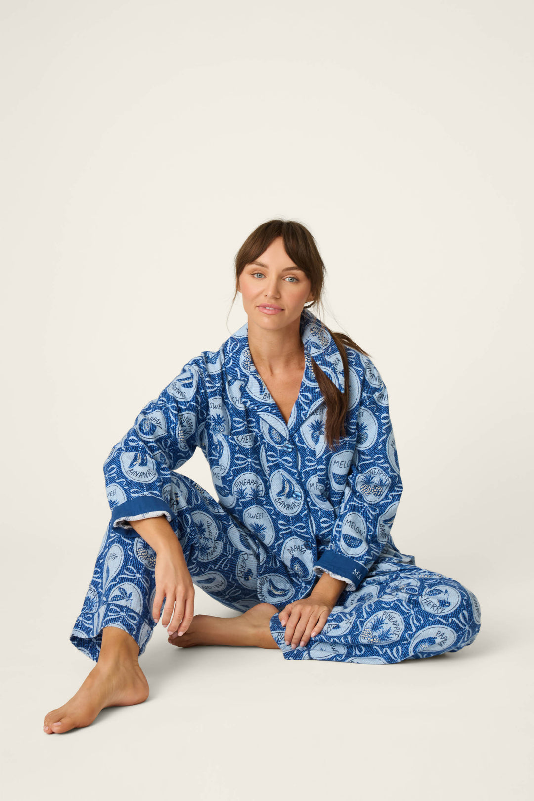 A woman sits on the floor against a plain light background, wearing the P.J. Salvage Fruit Medley Print 3-Piece Flannel PJ Set with one leg bent and her arm resting on her knee.