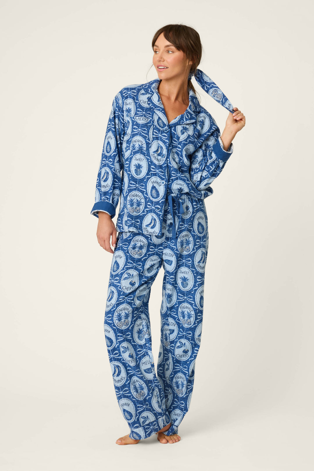 A woman stands barefoot and smiling against a plain light background, wearing the P.J. Salvage Fruit Medley Print 3-Piece Flannel PJ Set and holding the matching hair tie in her right hand.