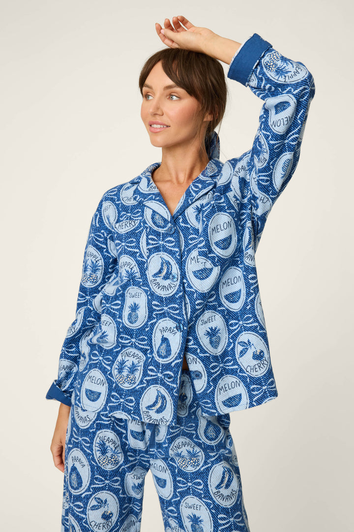 A woman stands against a light background, smiling and raising one arm to touch her head while wearing the P.J. Salvage Fruit Medley Print 3-Piece Flannel PJ Set featuring playful prints of melon, pineapple, and cherry.