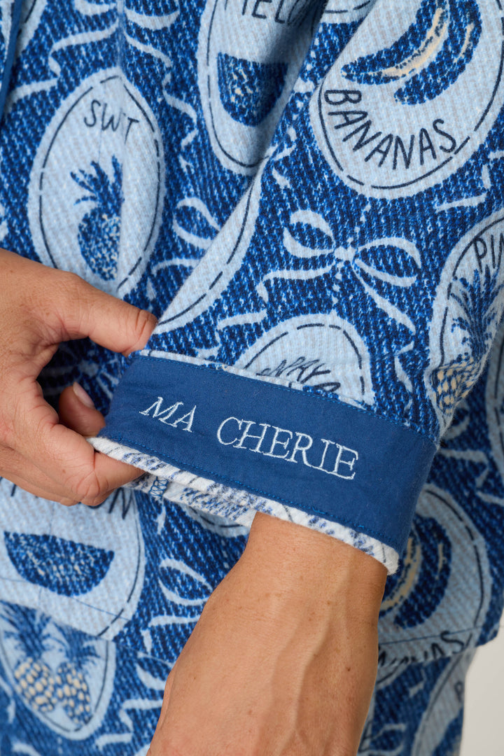 A hand holds the cuff of a blue patterned sleeve from the P.J. Salvage Fruit Medley Print 3-Piece Flannel PJ Set, featuring "MA CHERIE" embroidered in white thread and adorned with circular fruit and text designs in various shades of blue.