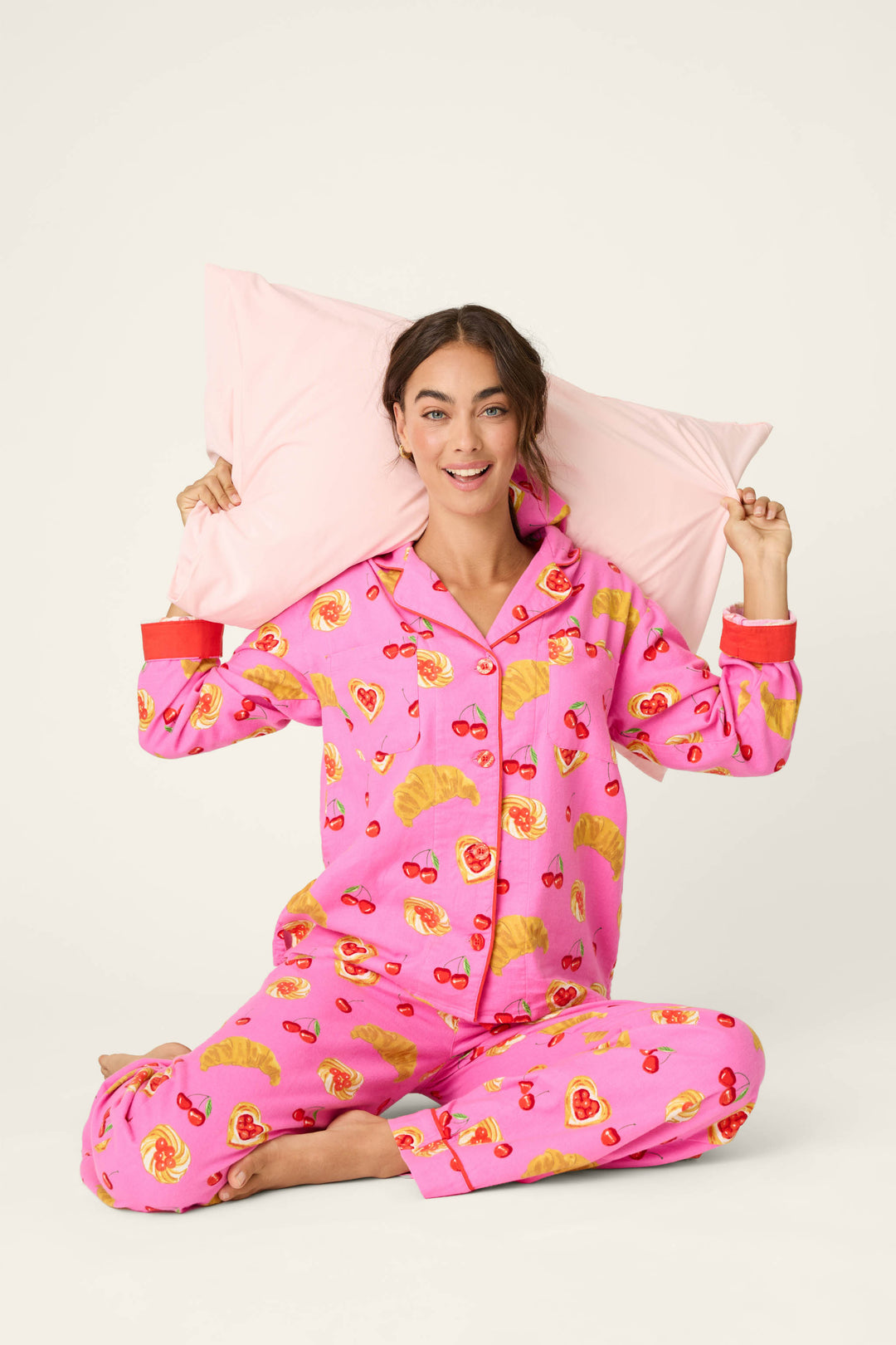 A woman smiles while sitting cross-legged on the floor against a plain background, holding a pink pillow behind her head and wearing the P.J. Salvage Breakfast Pastry Print 3-Piece Flannel PJ Set in bright pink.