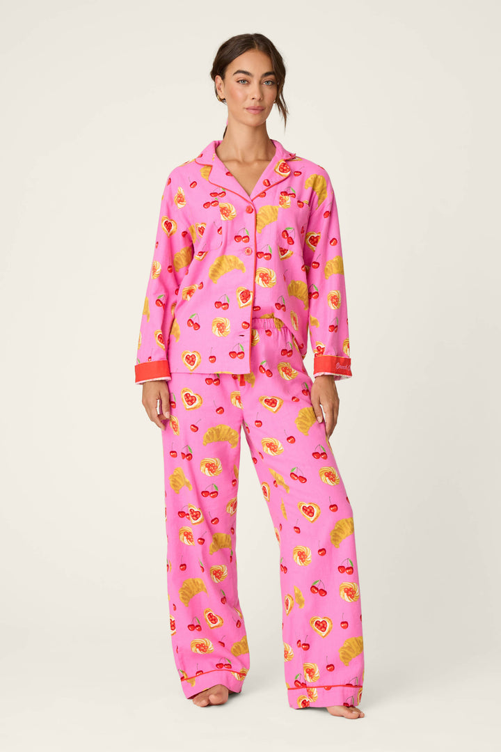 A person stands barefoot against a plain white background wearing the P.J. Salvage Breakfast Pastry Print 3-Piece Flannel PJ Set, which features a bright pink color with croissant and heart patterns and red accents on the cuffs.
