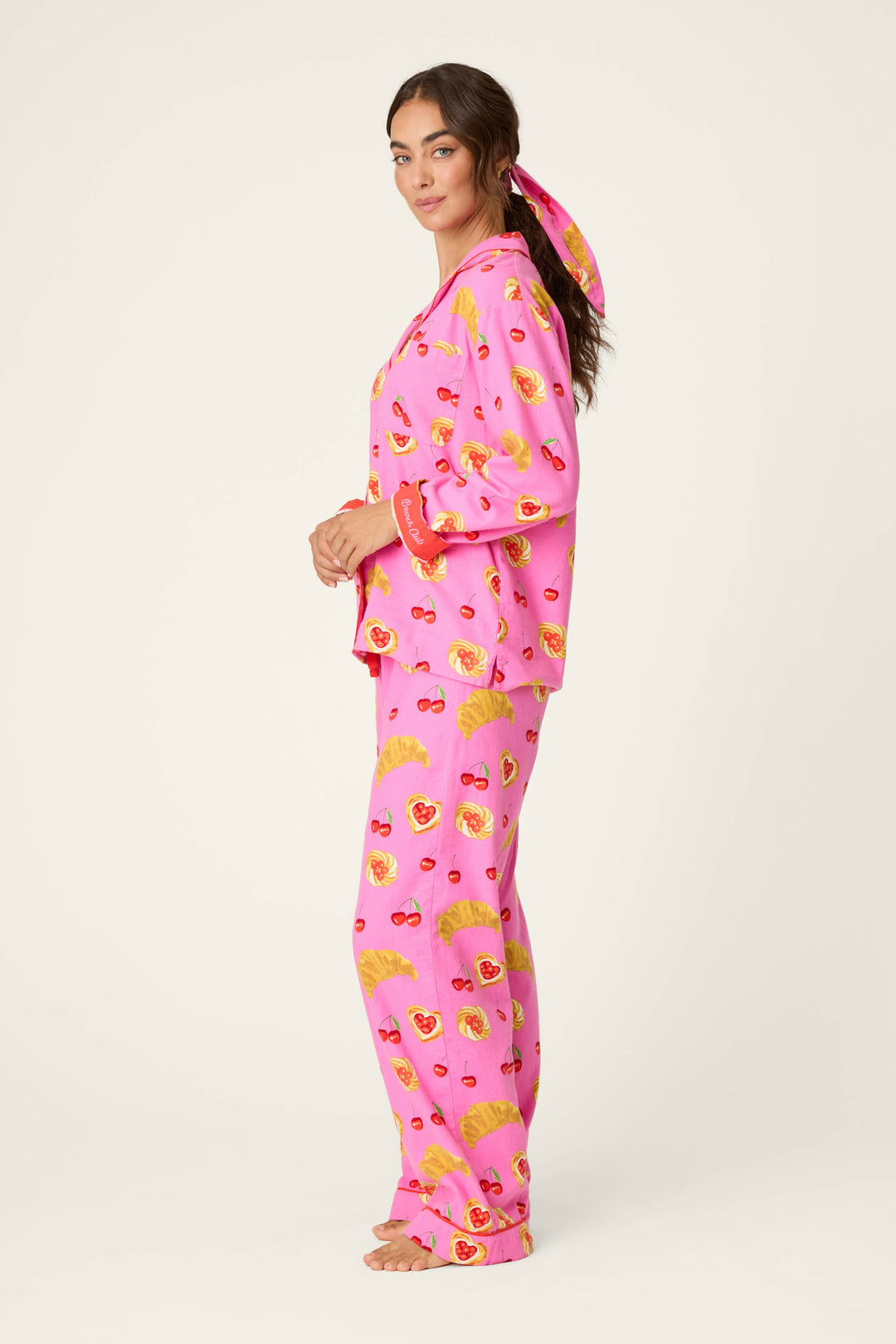 A woman stands in profile, dressed in the P.J. Salvage Breakfast Pastry Print 3-Piece Flannel PJ Set featuring bright pink fabric adorned with playful yellow and red pastry patterns. Her hair is pulled back with a coordinating pink scarf, set against a plain, off-white background.