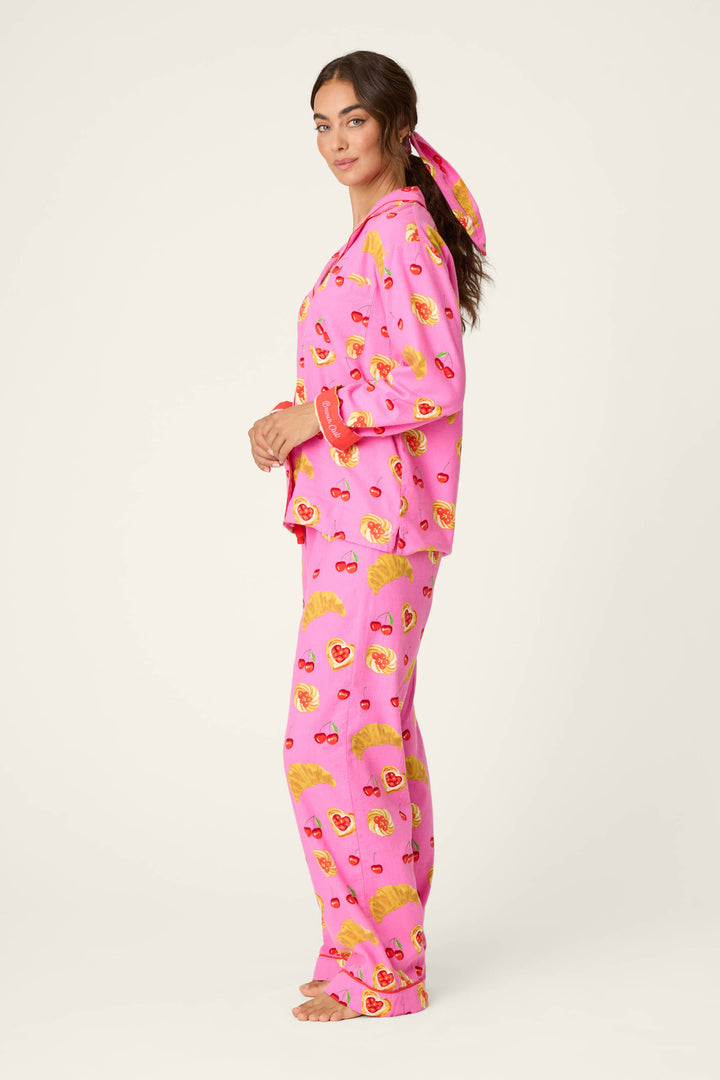 A woman stands in profile, dressed in the P.J. Salvage Breakfast Pastry Print 3-Piece Flannel PJ Set featuring bright pink fabric adorned with playful yellow and red pastry patterns. Her hair is pulled back with a coordinating pink scarf, set against a plain, off-white background.