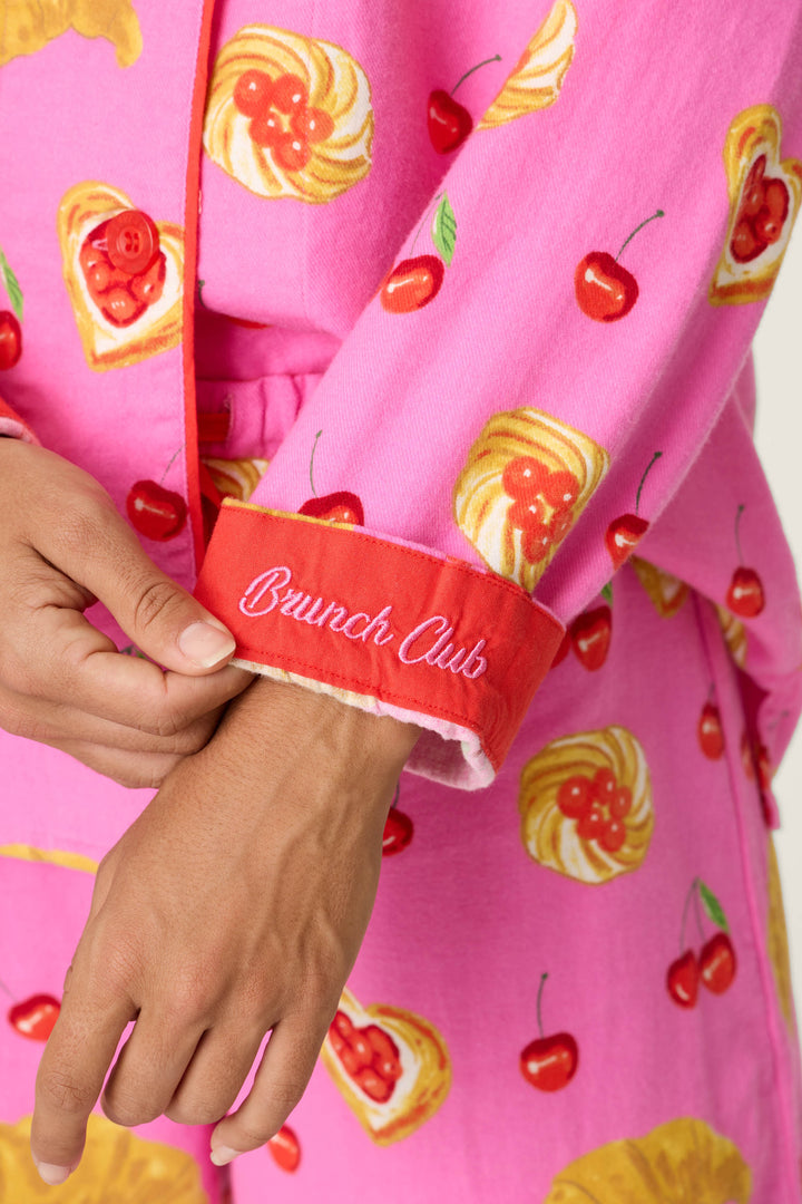 A person dressed in the P.J. Salvage Breakfast Pastry Print 3-Piece Flannel PJ Set, featuring pink pajamas with cherry and pastry designs, holds a sleeve with a red cuff embroidered "Brunch Club" in white script.
