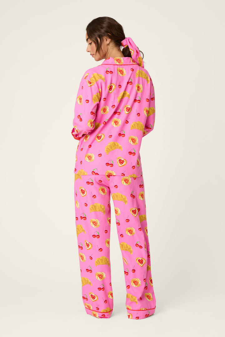 A woman stands facing away, wearing the P.J. Salvage Breakfast Pastry Print 3-Piece Flannel PJ Set, featuring pink pajamas patterned with red cherries, yellow fans, and hearts. Her brown hair is tied back with a matching pink scrunchie. The background is plain and light-colored.