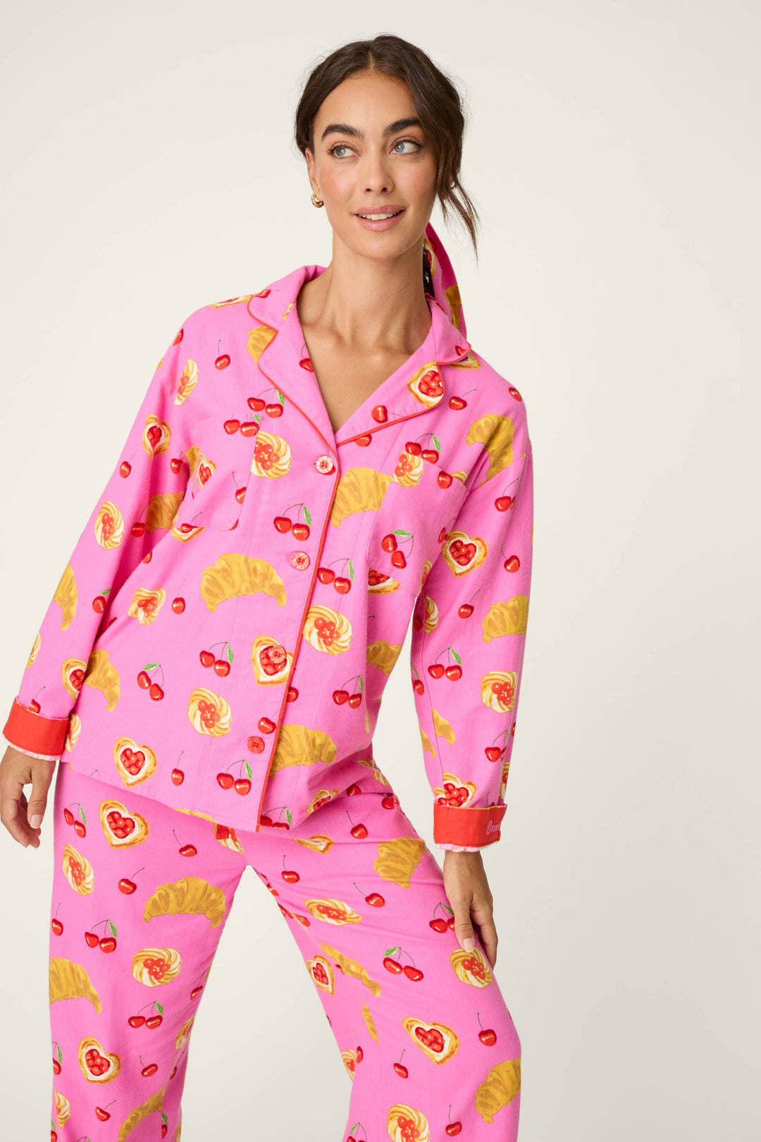 A woman models the P.J. Salvage Breakfast Pastry Print 3-Piece Flannel PJ Set, featuring a cheerful pattern of croissants, cherries, and heart-shaped pastries in bright pink. She smiles while standing against a simple light background.