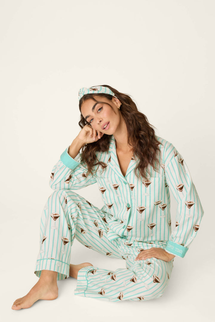 A woman sits barefoot on the floor, her head resting on her hand, wearing the P.J. Salvage Espresso Martini Print 3-Piece Long Sleeve Flannel PJ Set with a matching headband. Her long, wavy hair frames her face, and the light-colored background is plain.