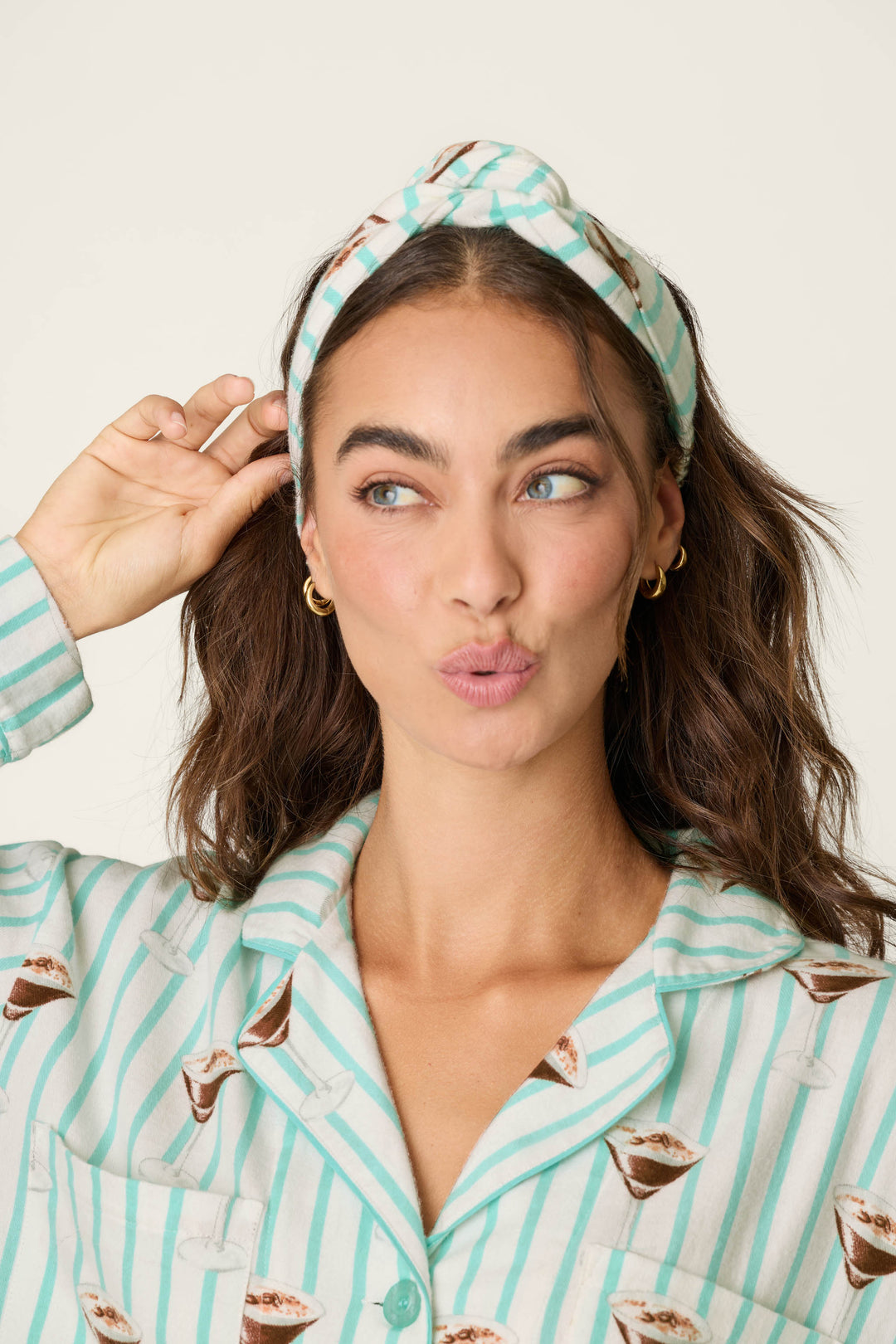 A woman with long brown hair wears the P.J. Salvage Espresso Martini Print 3-Piece Long Sleeve Flannel PJ Set, including a matching headband, and makes a playful pouty face with raised eyebrows against a plain backdrop.