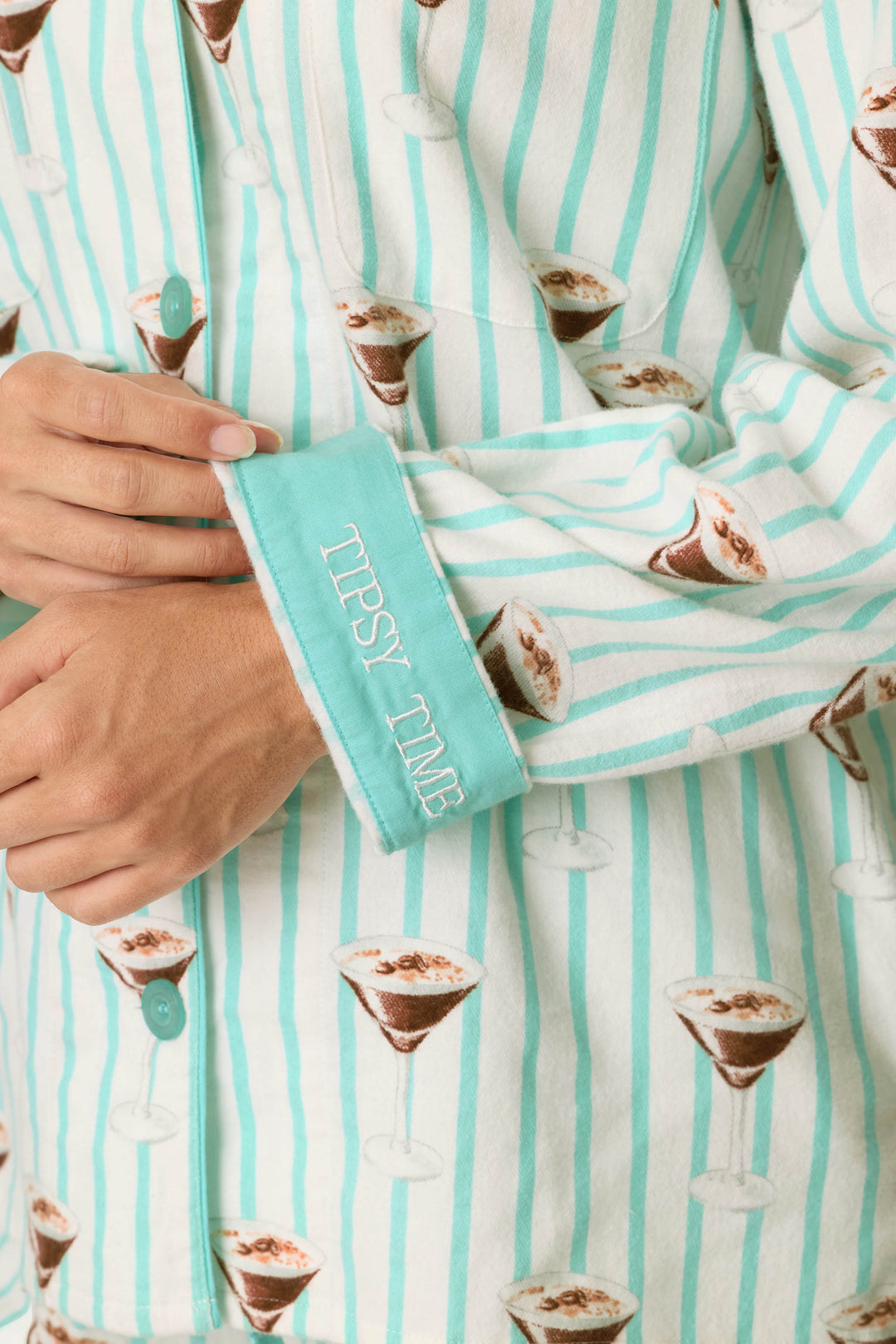 A person wearing the P.J. Salvage Espresso Martini Print 3-Piece Long Sleeve Flannel PJ Set, featuring blue stripes and martini glass prints, with the cuff flipped to reveal embroidered text reading “TIPSY TIME.”.