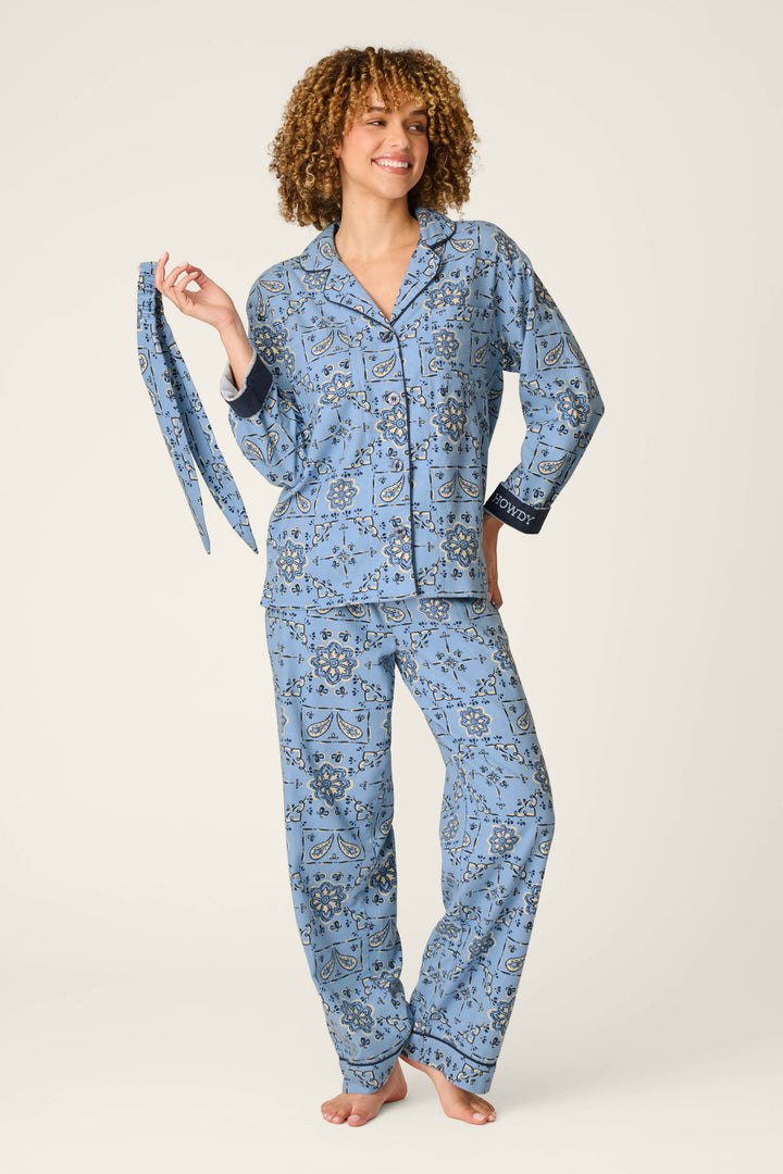 A person stands barefoot and smiling against a plain light background, wearing the P.J. Salvage Classic Paisley Bandana Print 3-Piece Long Sleeve Flannel PJ Set and holding the matching belt in their left hand.