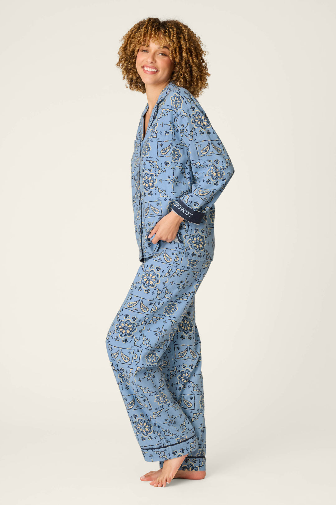 A smiling person with curly hair stands barefoot on one foot against a plain light background, wearing the P.J. Salvage Classic Paisley Bandana Print 3-Piece Long Sleeve Flannel PJ Set.