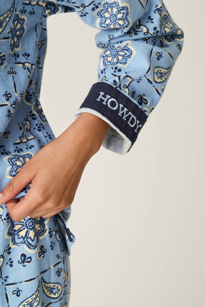 A person wearing the P.J. Salvage Classic Paisley Bandana Print 3-Piece Long Sleeve Flannel PJ Set, featuring blue patterned pajamas with a sleeve cuff embroidered with “HOWDY” in white letters.