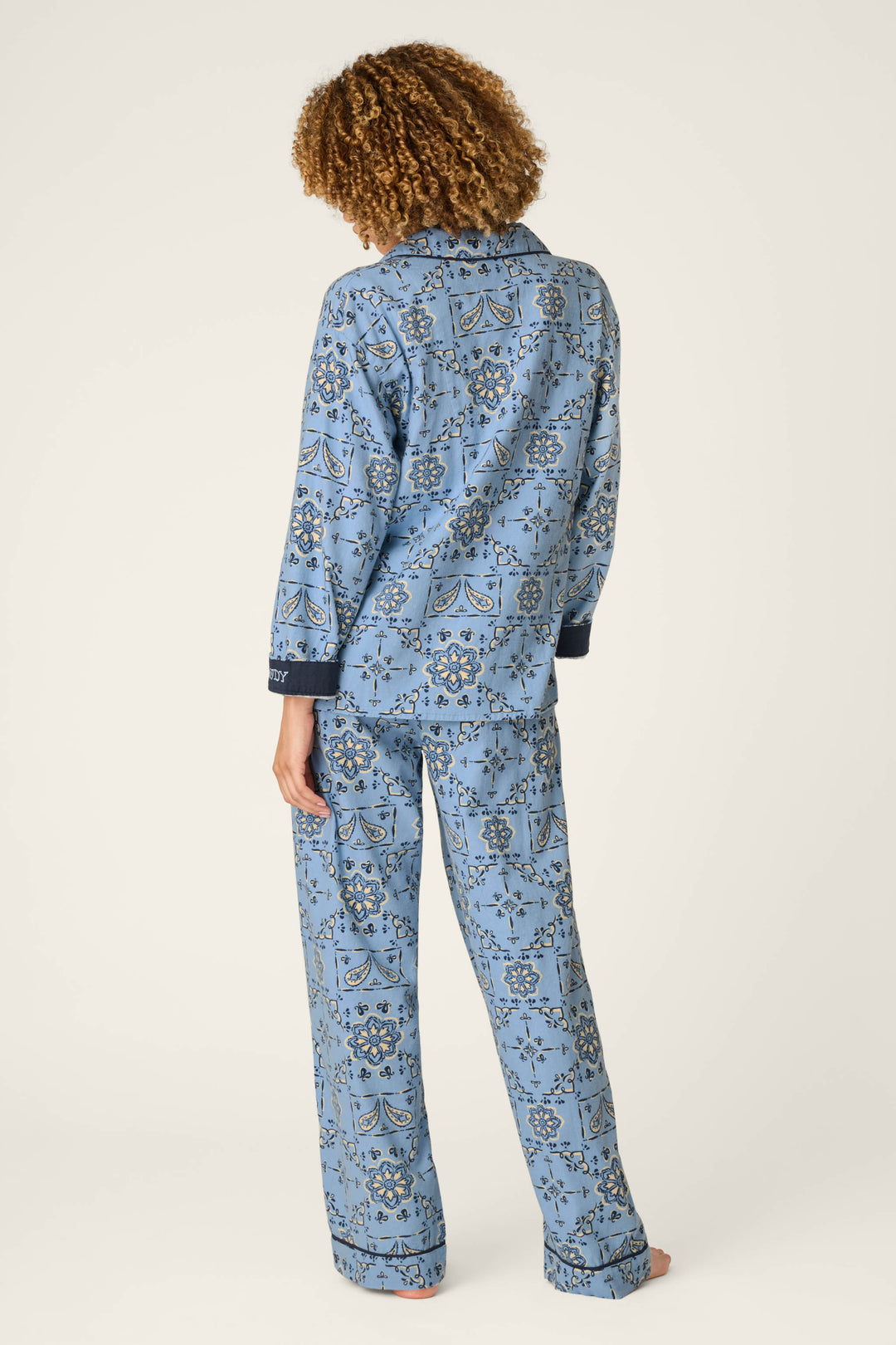 A person with curly hair stands barefoot, facing away, wearing the P.J. Salvage Classic Paisley Bandana Print 3-Piece Long Sleeve Flannel PJ Set against a plain light background.