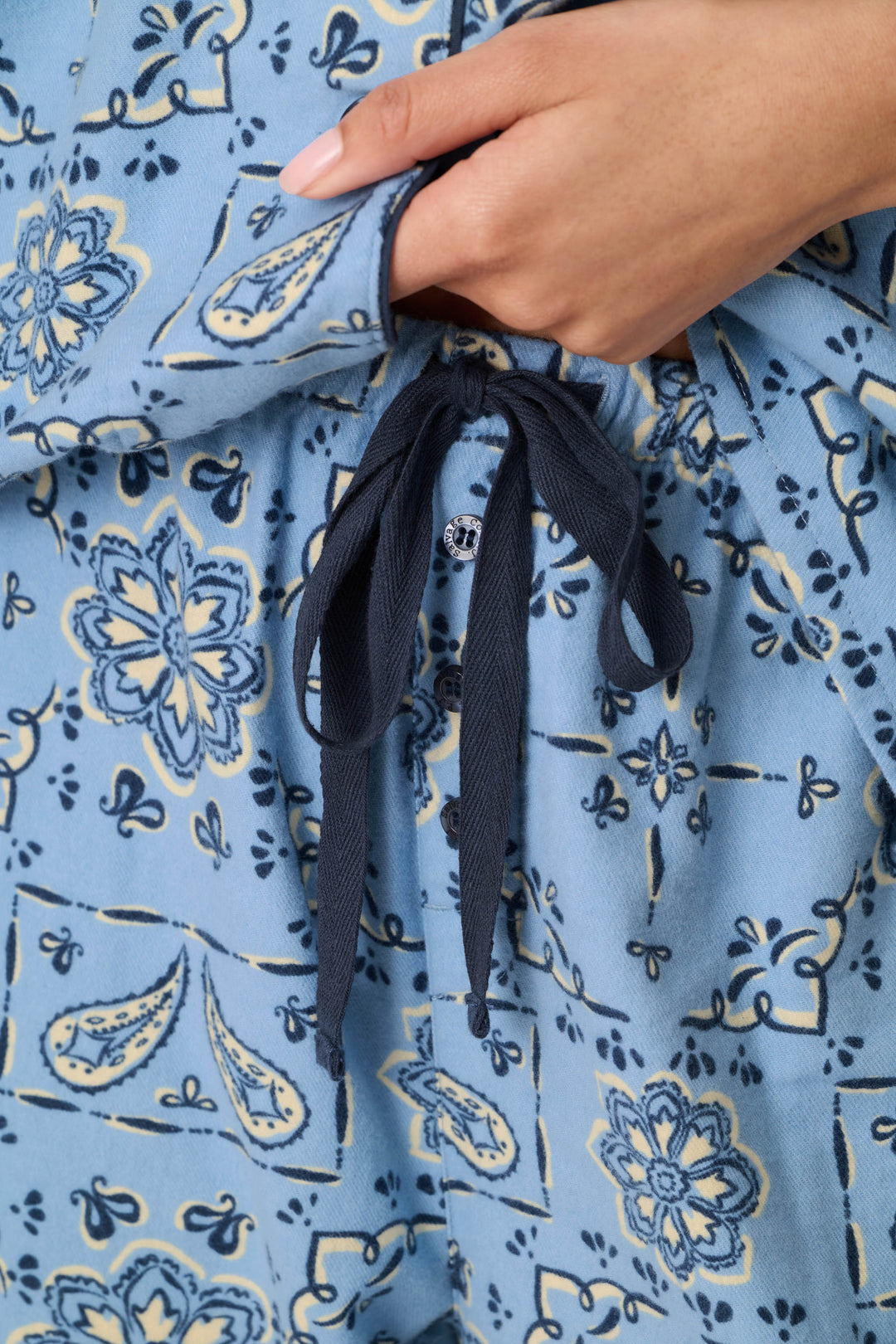 A close-up of a hand adjusting the navy blue drawstring on P.J. Salvage’s Classic Paisley Bandana Print 3-Piece Long Sleeve Flannel PJ Set, featuring light blue pants with a yellow and blue floral pattern.