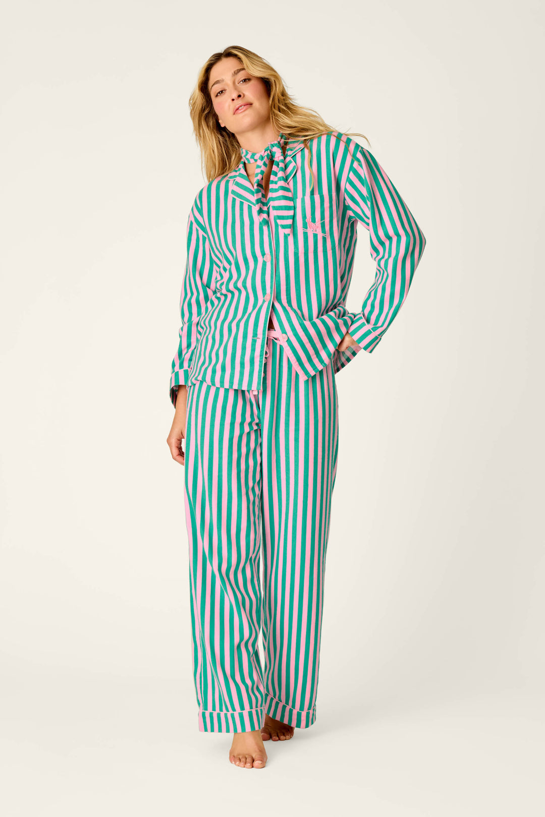 A woman stands barefoot against a plain light background, wearing the P.J. Salvage Ski Bunny Icon 3-Piece Long Sleeve Flannel PJ Set—loose-fitting pajamas featuring vertical pink, green, and white stripes.
