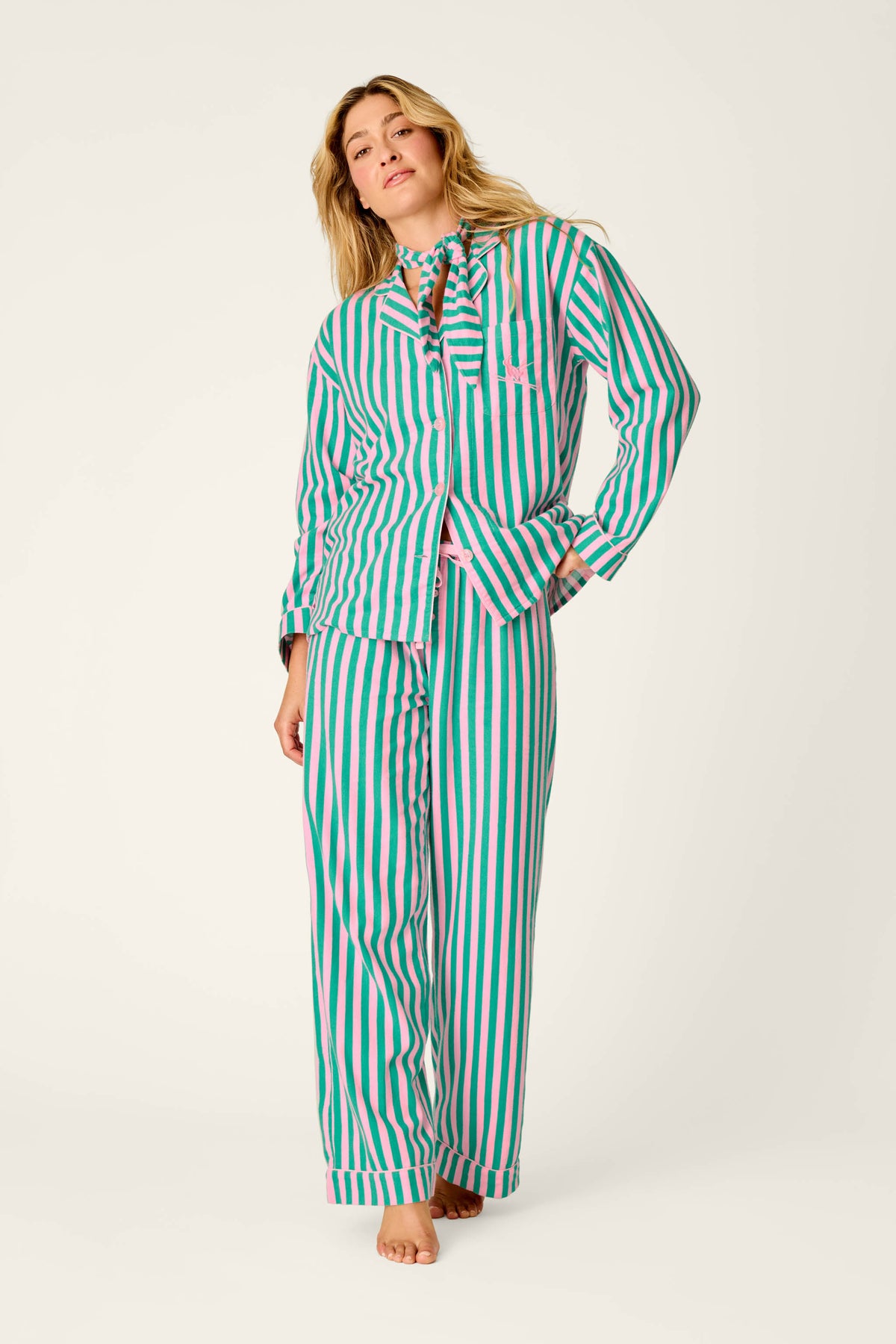 A woman stands barefoot against a plain light background, wearing the P.J. Salvage Ski Bunny Icon 3-Piece Long Sleeve Flannel PJ Set—loose-fitting pajamas featuring vertical pink, green, and white stripes.