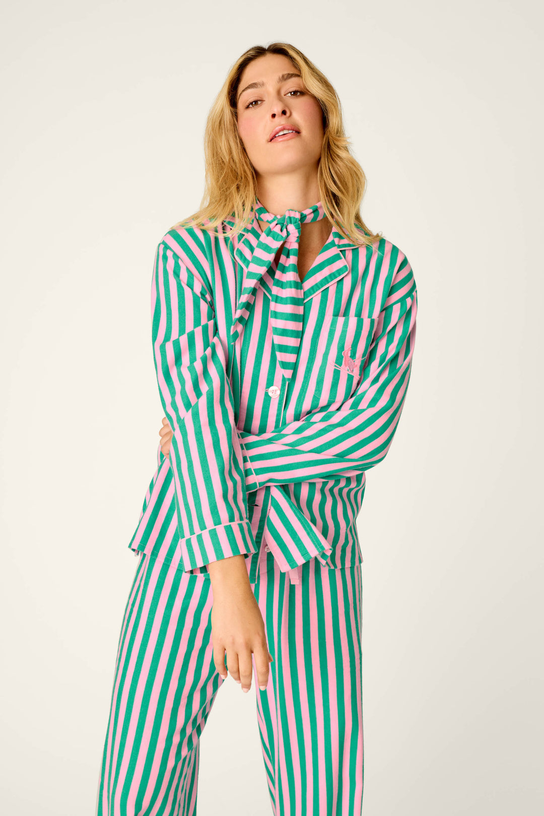 A woman with long blonde hair wears the P.J. Salvage Ski Bunny Icon 3-Piece Long Sleeve Flannel PJ Set, featuring matching green, pink, and white vertical stripes, as she stands with one arm across her body against a plain light background.