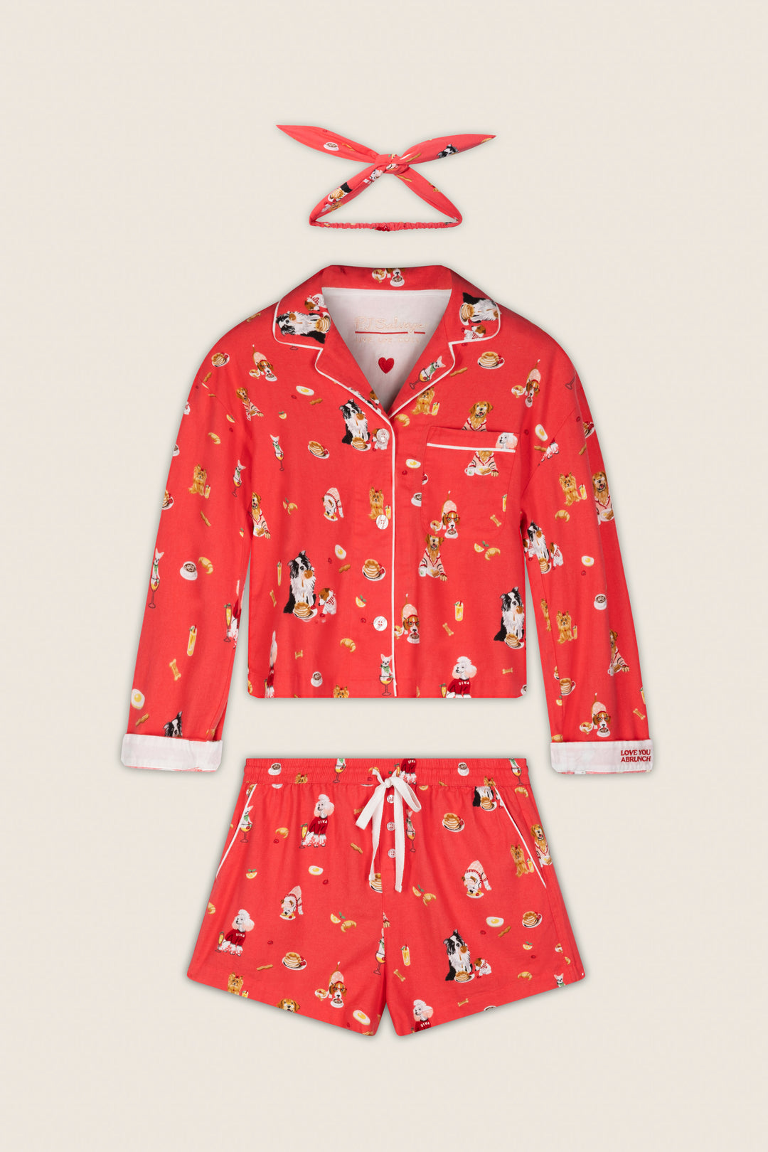 The P.J. Salvage Dogs Who Brunch Print 3-Piece Flannel PJ Set, featuring a collared red top with white trim and a front pocket, matching shorts, and a headband—all adorned with festive cartoon dog prints—is displayed flat on a light background.