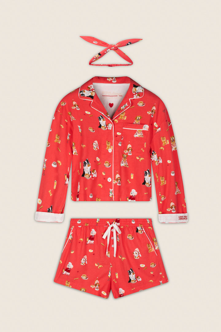 The P.J. Salvage Dogs Who Brunch Print 3-Piece Flannel PJ Set, featuring a collared red top with white trim and a front pocket, matching shorts, and a headband—all adorned with festive cartoon dog prints—is displayed flat on a light background.