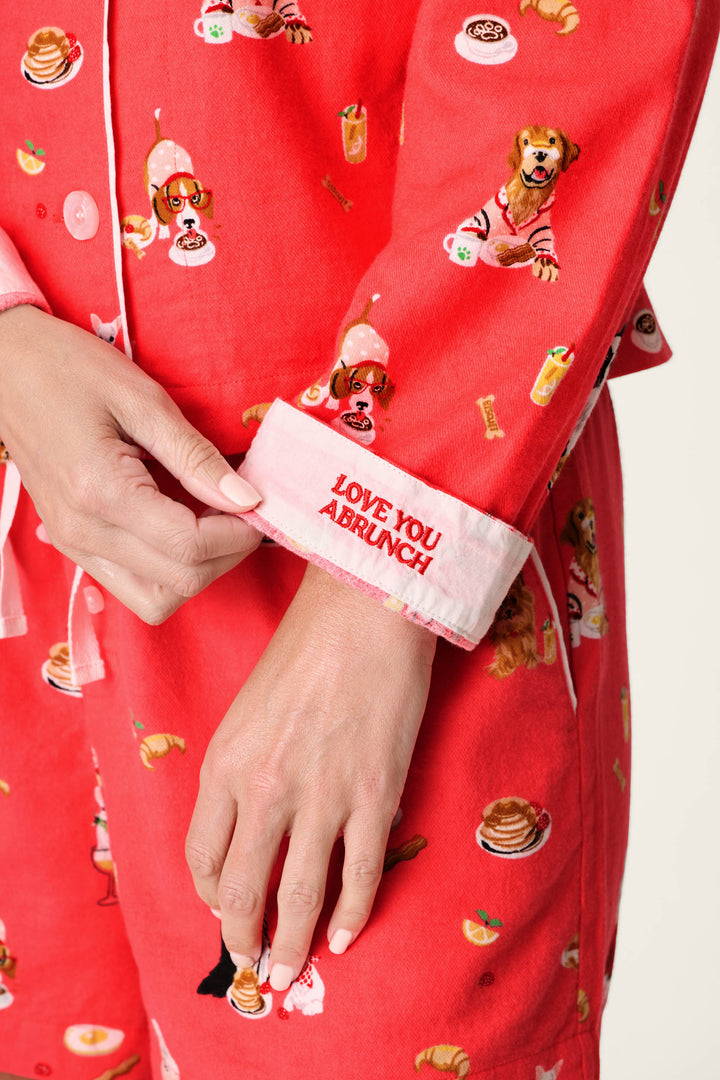 Wearing the P.J. Salvage Dogs Who Brunch Print 3-Piece Flannel PJ Set, which features playful breakfast food and dog prints on red flannel, a person turns up a cuff to reveal embroidered text that reads: LOVE YOU A BRUNCH. Only their lower torso and hand are shown.
