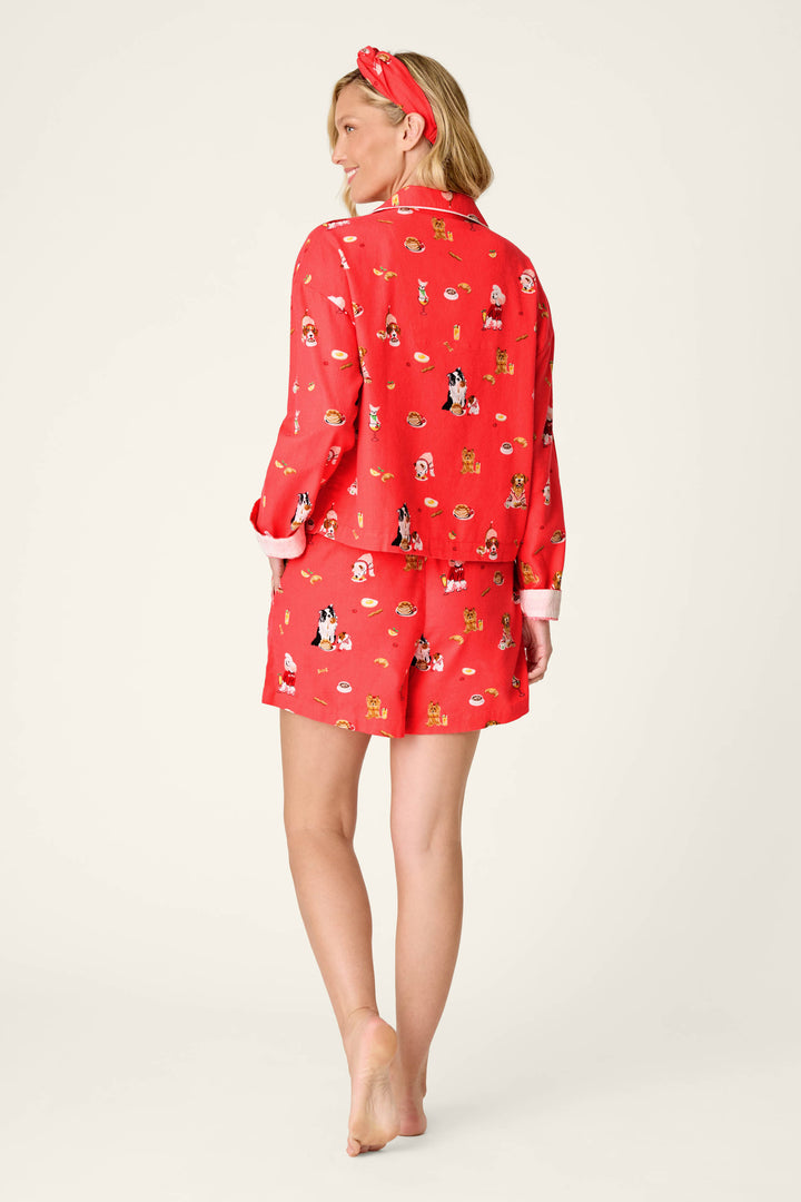 A woman stands barefoot, facing away, wearing the P.J. Salvage Dogs Who Brunch Print 3-Piece Flannel PJ Set in red, featuring small dog illustrations. Her blonde hair is styled in loose waves and she accessorizes with a matching red headband. The background is plain and light-colored.