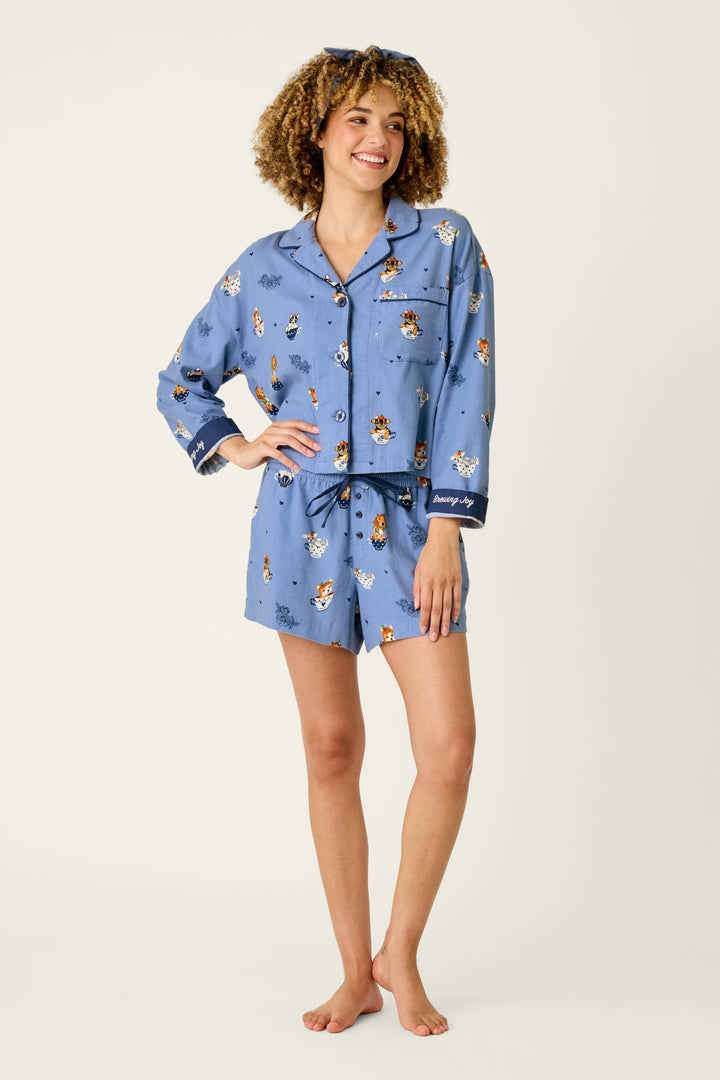 A smiling woman with curly hair models the P.J. Salvage Teacup Pups 3-Piece Short Flannel PJ Set, a blue pajama set with cartoon dog prints, featuring a long-sleeve button-up top and matching shorts, as she stands barefoot against a plain light background.