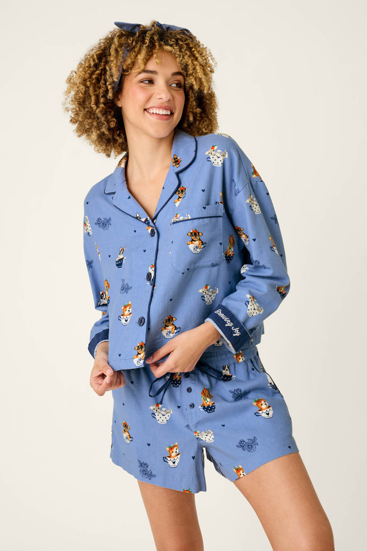 A smiling woman with curly hair models the P.J. Salvage Teacup Pups 3-Piece Short Flannel PJ Set, featuring playful cartoon dog prints and a coordinating headband, standing against a plain background.