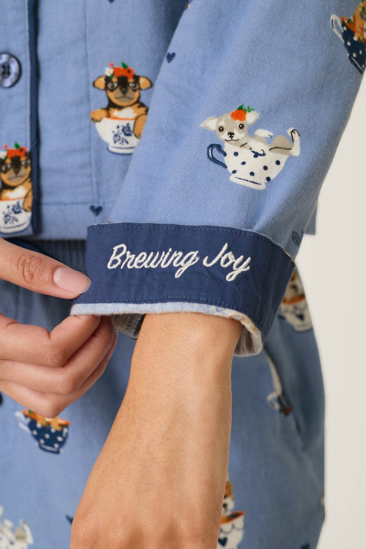Wearing the P.J. Salvage Teacup Pups 3-Piece Short Flannel PJ Set, a person adjusts the cuff of their blue pajamas, which are adorned with dogs in teacups and finished with “Brewing Joy” embroidered in white script.