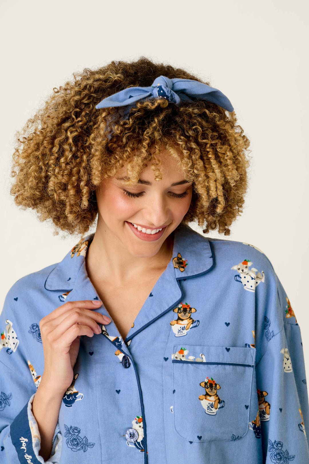 A woman with curly hair wearing a blue headband smiles while looking down, dressed in the P.J. Salvage Teacup Pups 3-Piece Short Flannel PJ Set featuring playful cartoon dog and heart prints.