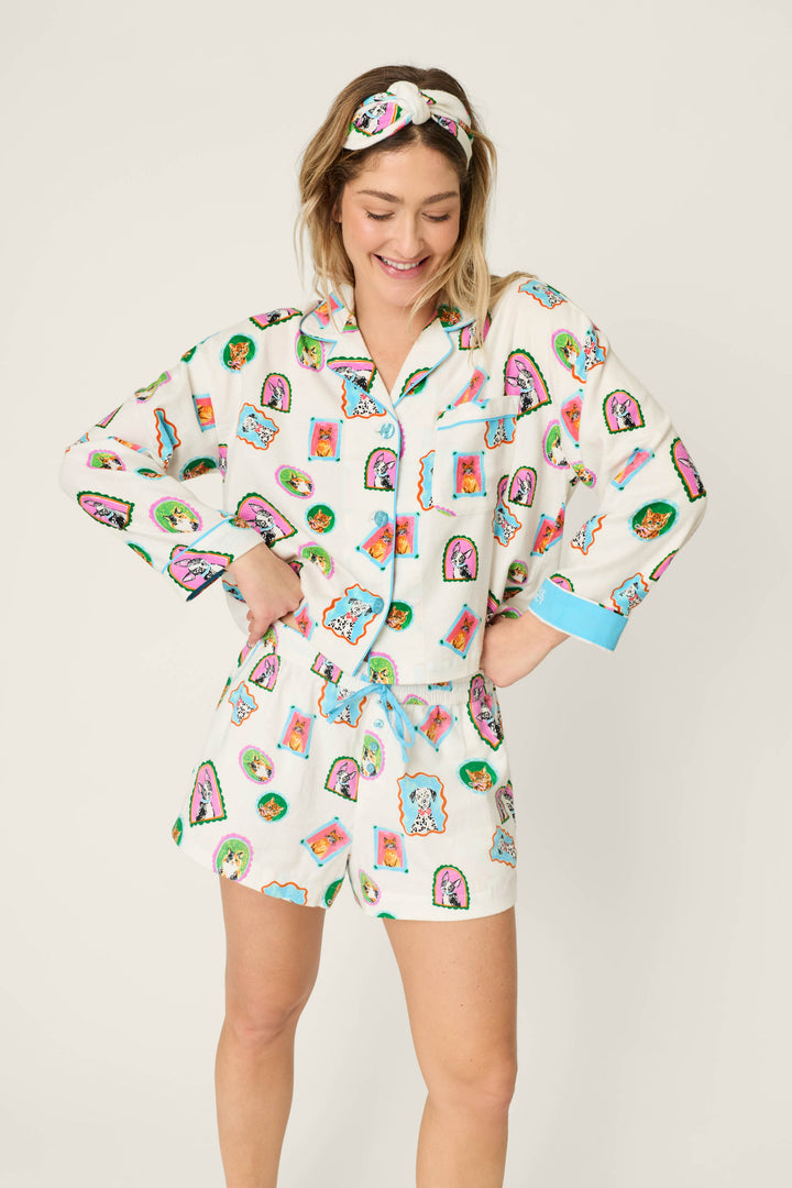 A smiling woman stands with her hands on her hips against a plain background, wearing the P.J. Salvage Picture Purrfect Pet Portraits 3-Piece Short Flannel PJ Set and a matching sleep mask decorated with colorful patterns.