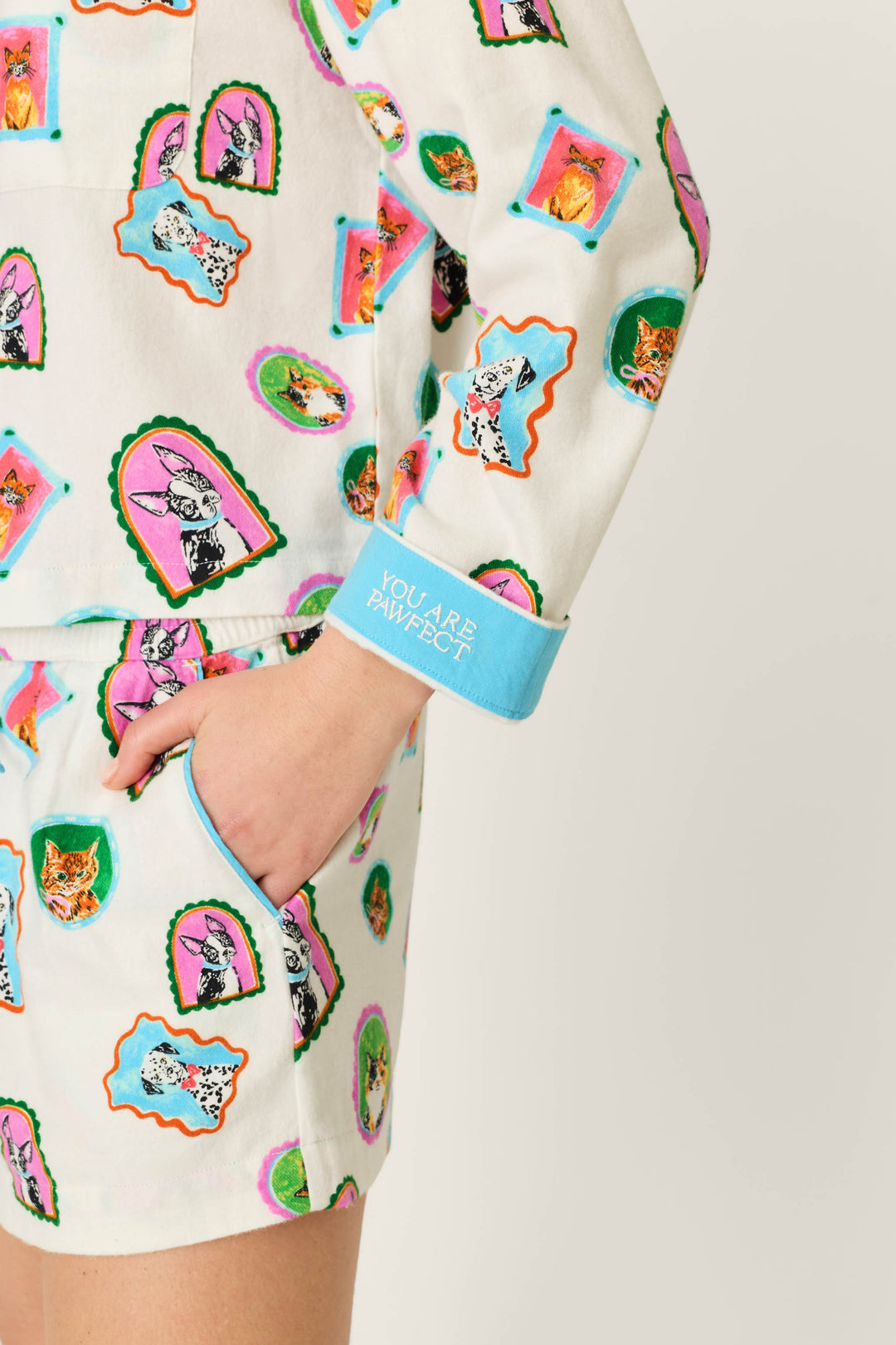 A person is wearing the P.J. Salvage Picture Purrfect Pet Portraits 3-Piece Short Flannel PJ Set, featuring a white base with playful cat and dog prints. Their left hand rests in a pocket, and the light blue cuff reads 'YOU ARE PAWFECT.' The background is plain and light-colored.