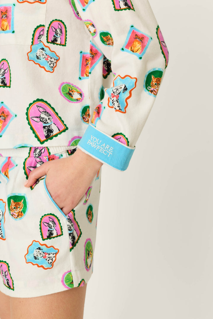 A person is wearing the P.J. Salvage Picture Purrfect Pet Portraits 3-Piece Short Flannel PJ Set, featuring a white base with playful cat and dog prints. Their left hand rests in a pocket, and the light blue cuff reads 'YOU ARE PAWFECT.' The background is plain and light-colored.