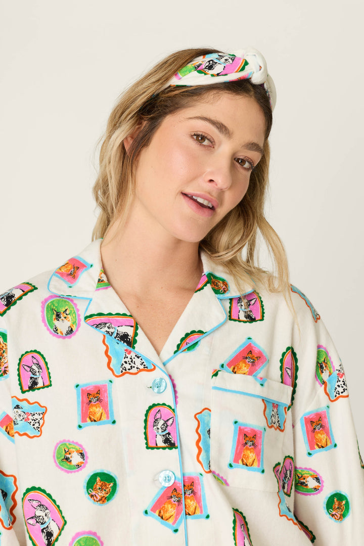 A woman wearing the P.J. Salvage Picture Purrfect Pet Portraits 3-Piece Short Flannel PJ Set, featuring vibrant cat and dog prints and a coordinating headband, smiles softly at the camera against a simple, light background.