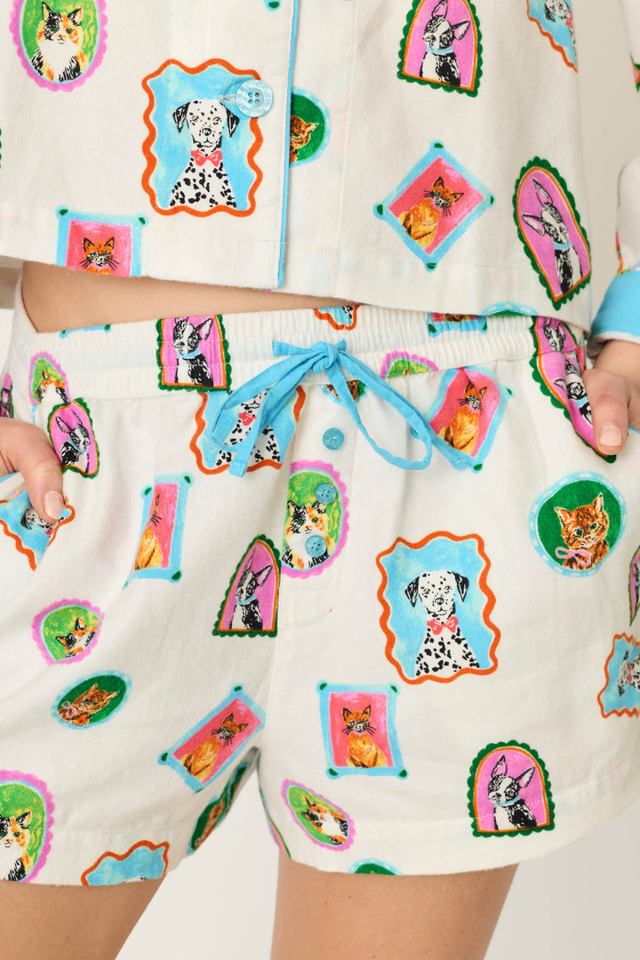 A person wearing the P.J. Salvage Picture Purrfect Pet Portraits 3-Piece Short Flannel PJ Set, featuring a matching pajama set adorned with colorful cartoon portraits of dogs and cats; the shorts are secured with a blue drawstring, and only the lower torso and hands are visible.