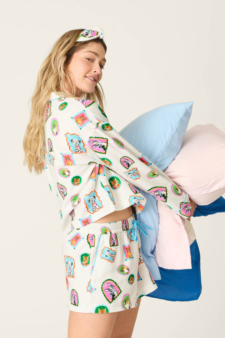 Wearing the P.J. Salvage Picture Purrfect Pet Portraits 3-Piece Short Flannel PJ Set and matching sleep mask, a woman playfully smiles while holding blue, pink, and white pillows against a plain light background.