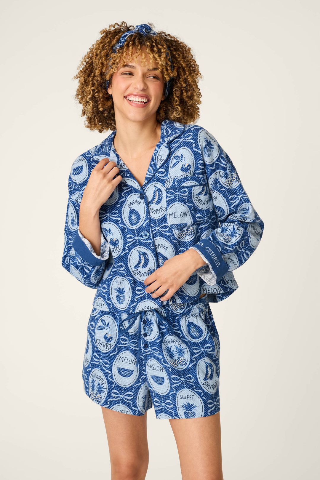 Wearing the P.J. Salvage Fruit Medley Print 3-Piece Short Flannel PJ Set, a person with curly hair smiles and stands with one hand on their shirt. The blue and white patterned pajama set, complete with a matching headband, features colorful fruits and playful words like "melon" and "sweet.
