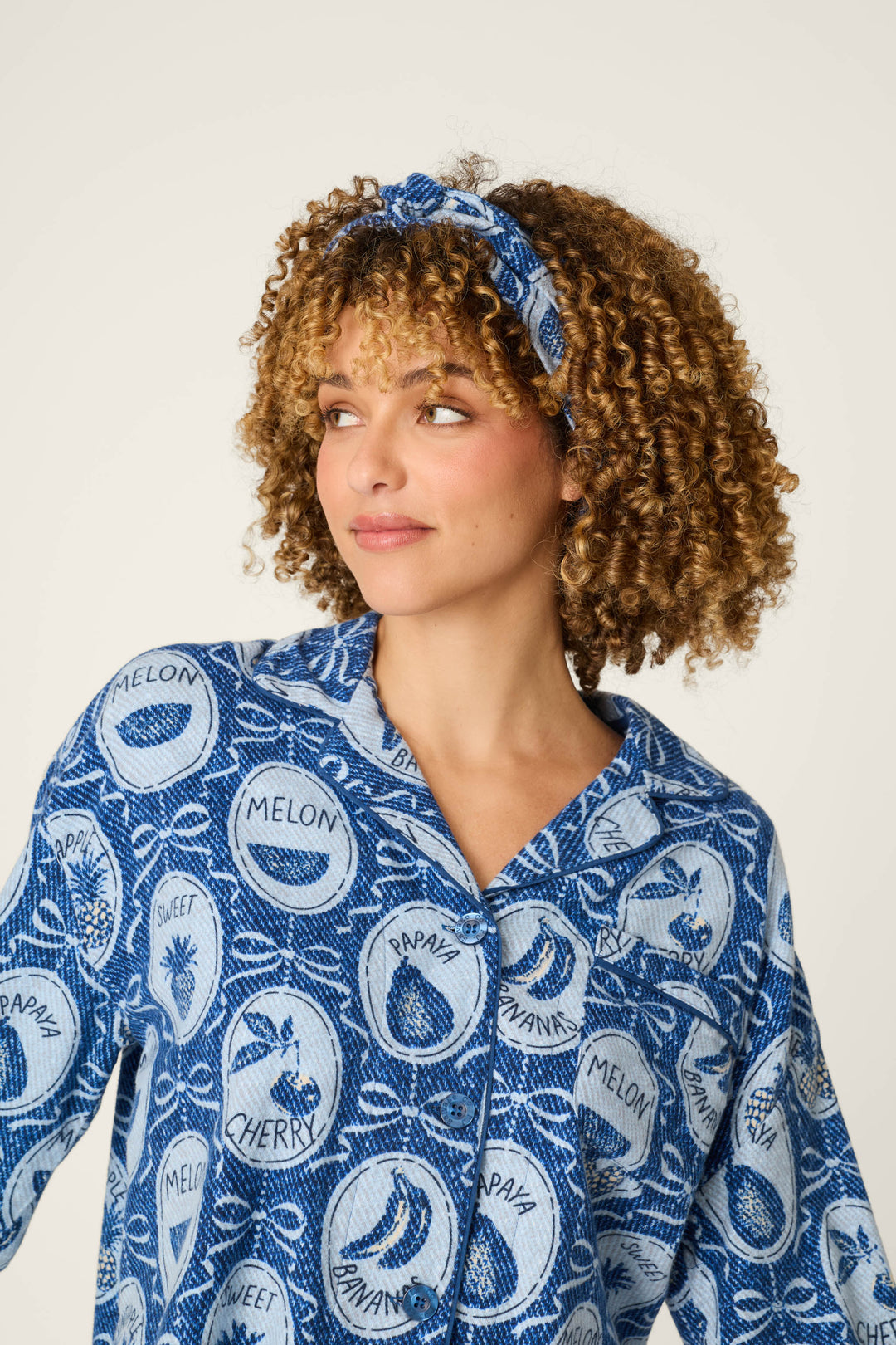 A woman with curly hair models the P.J. Salvage Fruit Medley Print 3-Piece Short Flannel PJ Set, featuring a matching blue patterned headband and shirt with fruit designs and text like “melon” and “cherry,” while looking to the side against a plain background.