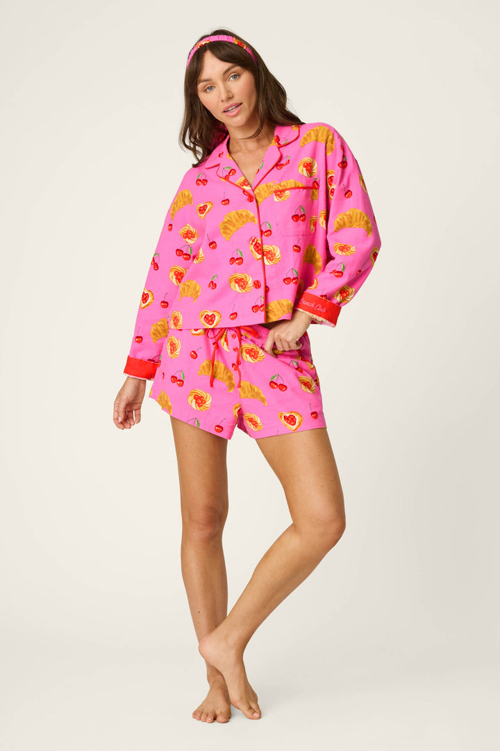 A woman stands barefoot on a plain background wearing the P.J. Salvage Breakfast Pastry Print 3-Piece Short Flannel PJ Set, featuring a matching pink long-sleeve button-up shirt, shorts, and headband with playful yellow and red breakfast pastry patterns.