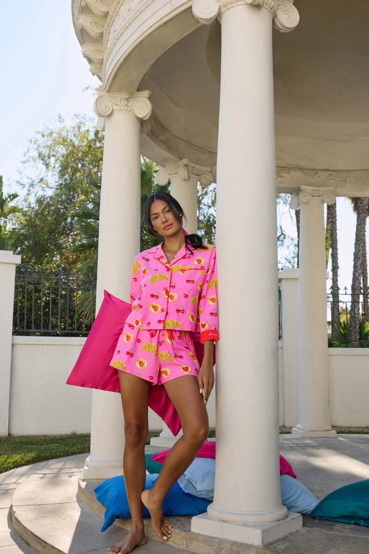 A woman stands barefoot by white columns, holding a matching pillow while wearing the P.J. Salvage Breakfast Pastry Print 3-Piece Short Flannel PJ Set. Colorful pillows are scattered on the ground, and trees can be seen in the background.