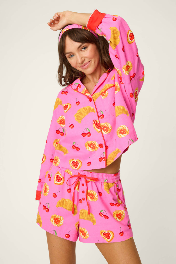 A woman smiles as she poses playfully in the P.J. Salvage Breakfast Pastry Print 3-Piece Short Flannel PJ Set, featuring bright pink pajamas with red cuffs adorned with pastries, cherries, and hearts. She completes the look with a matching headband, standing against a plain background.
