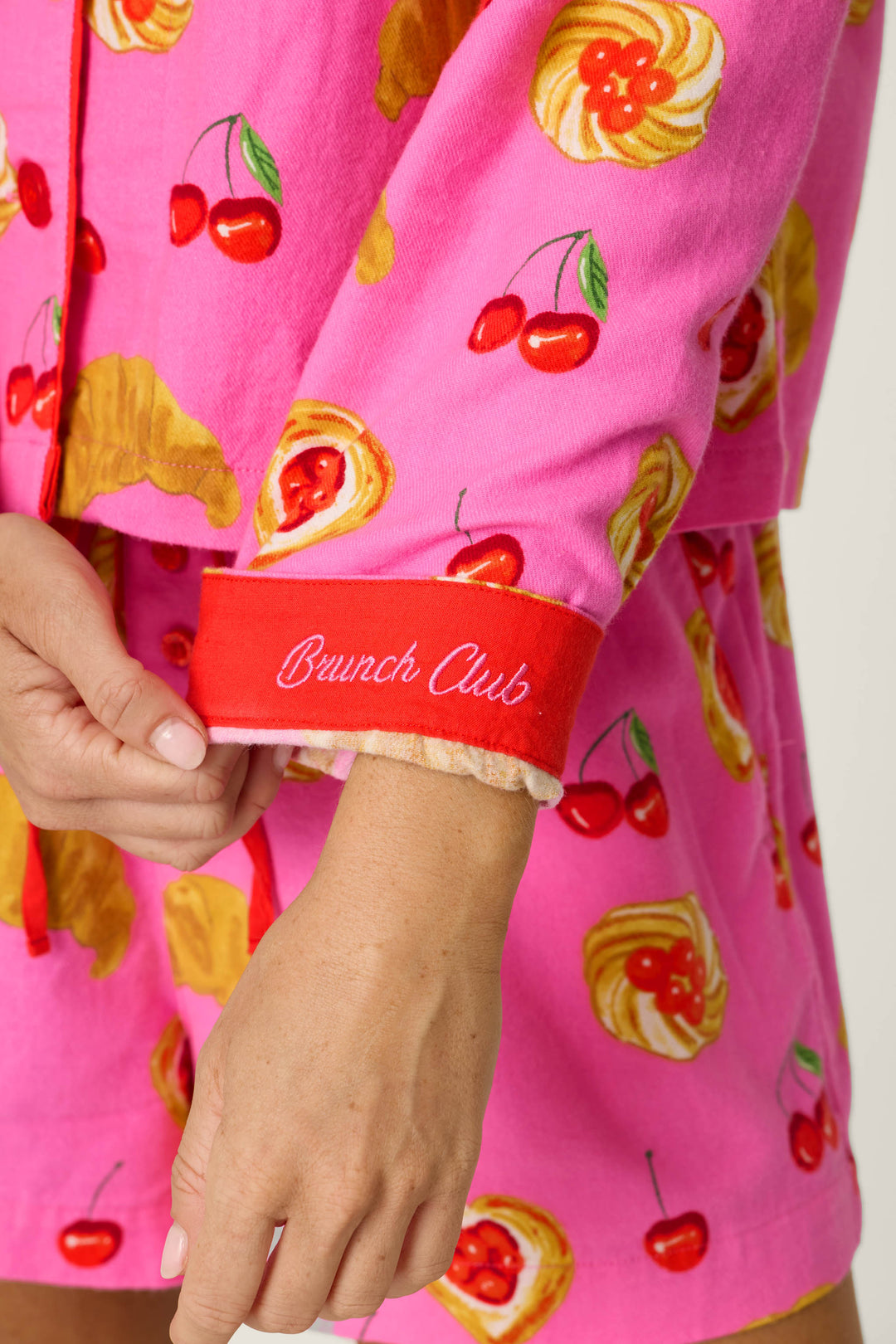 A person is wearing the P.J. Salvage Breakfast Pastry Print 3-Piece Short Flannel PJ Set, featuring vibrant pink pajamas with cherry and pastry designs. The visible red sleeve cuff is embroidered with "Brunch Club" in white cursive, showing only the hand and lower arm.