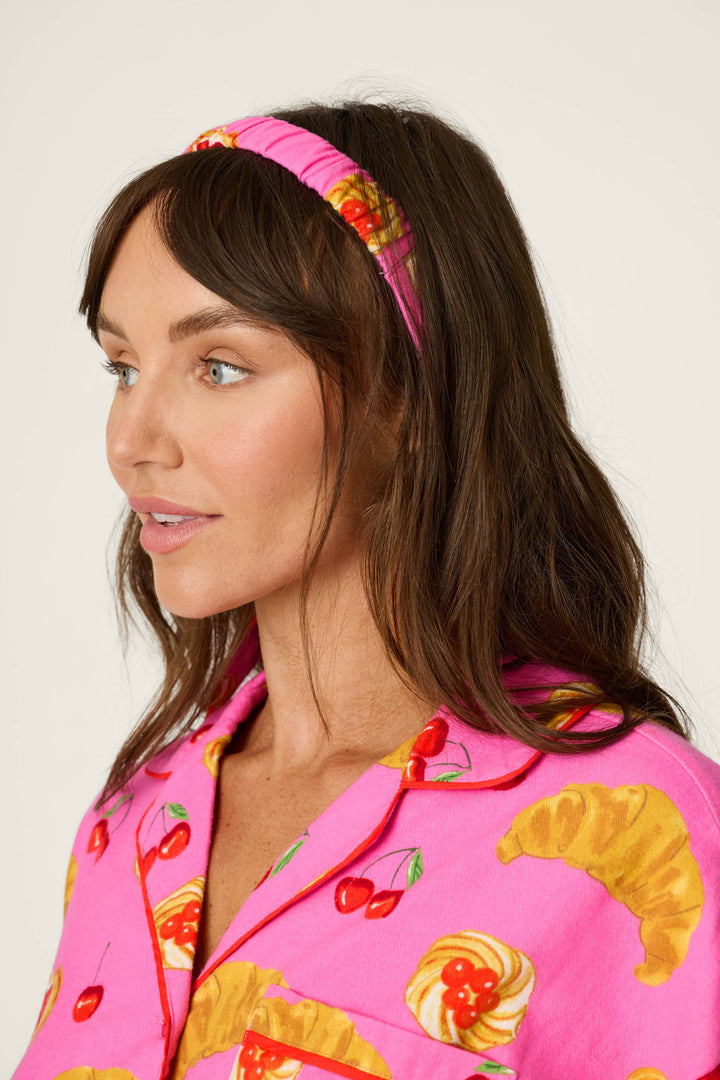 A woman with long brown hair, wearing P.J. Salvage’s Breakfast Pastry Print 3-Piece Short Flannel PJ Set featuring a pink headband and a pink shirt decorated with croissant, cherry, and pastry prints, looks to the left against a plain background.