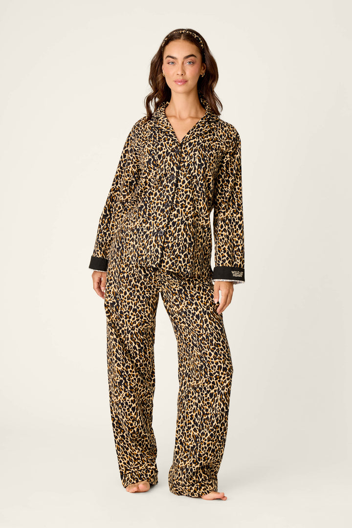 A woman stands barefoot wearing the P.J. Salvage Wild At Heart Leopard Print 3-Piece Flannel PJ Set, featuring a matching leopard print design with long sleeves and full-length pants. She has long, wavy hair styled with a headband and poses against a plain light background.