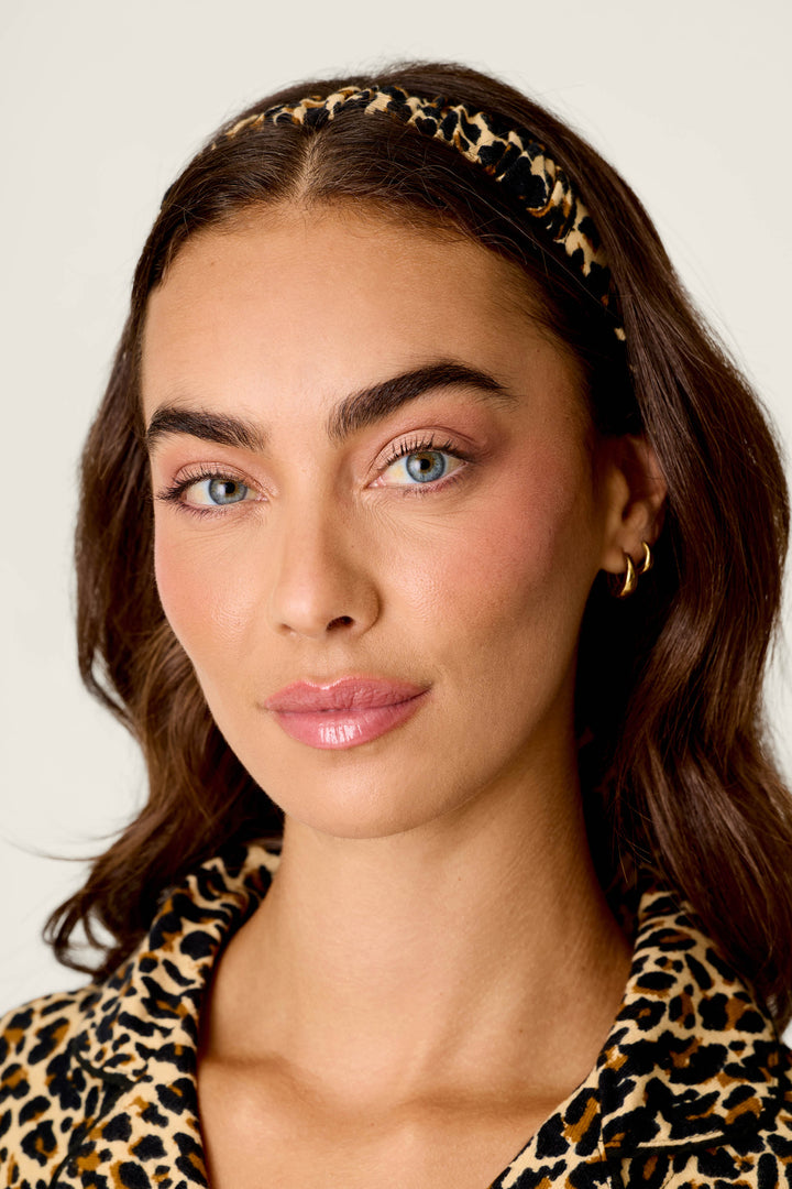 A woman with wavy brown hair and natural makeup, wearing the P.J. Salvage Wild At Heart Leopard Print 3-Piece Flannel PJ Set and gold hoop earrings, stands in front of a plain light background with a neutral expression.